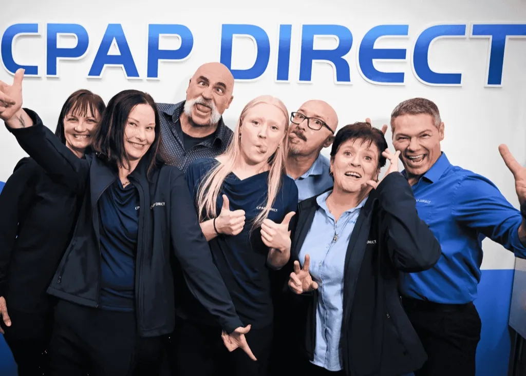 The CPAP Direct team crowded together in navy and blue uniform shirts, grinning and pulling faces under a large blue CPAP DIRECT sign